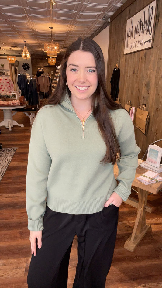 Woman in a green sweater and black pants standing in a store with wooden floors and clothing racks.