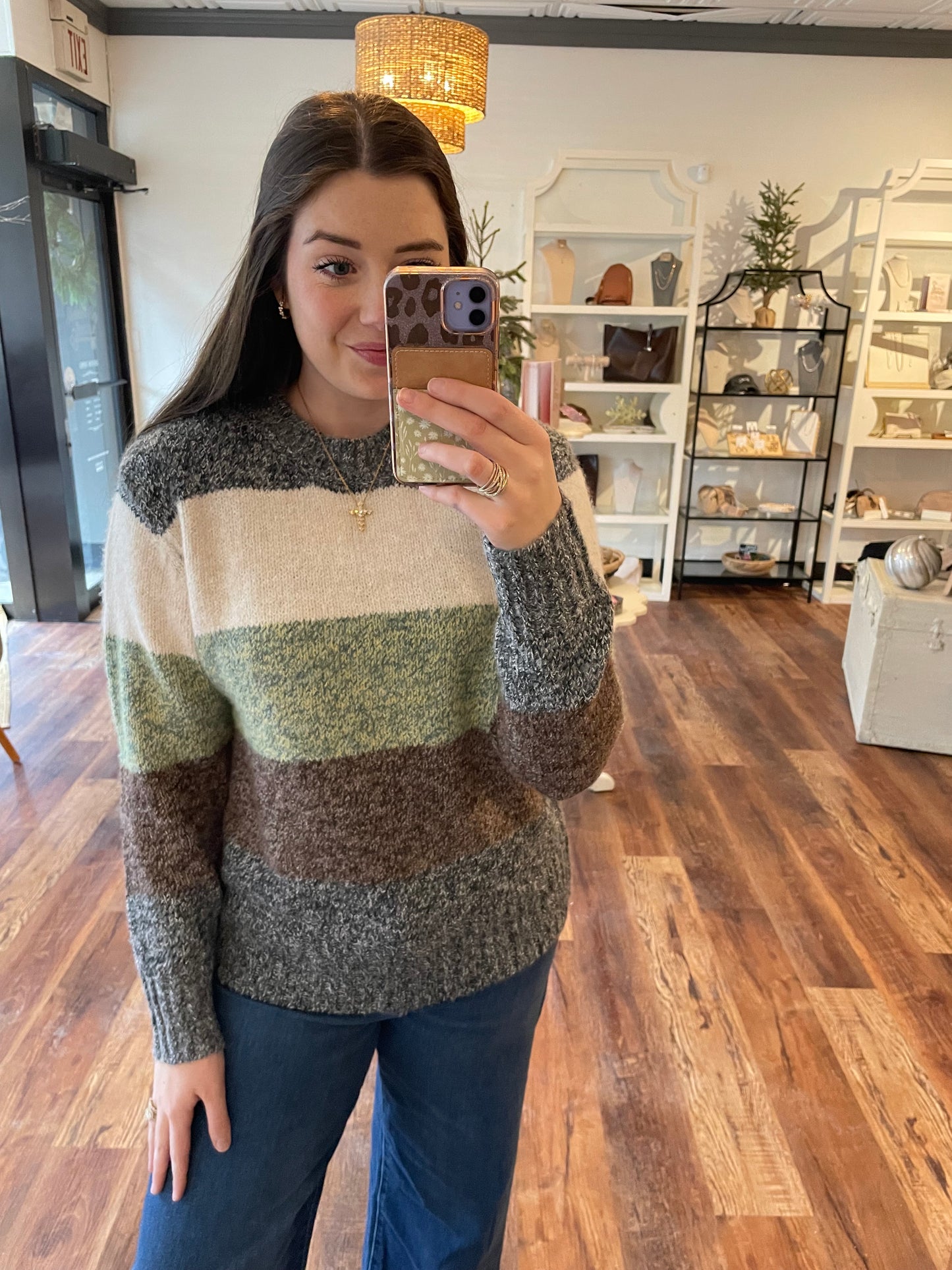 Person wearing a striped sweater taking a mirror selfie in a room with wooden flooring and decorative items.